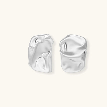 Pair of sculptural silver ear cuffs with organic fluid shapes and polished finish against cream background.