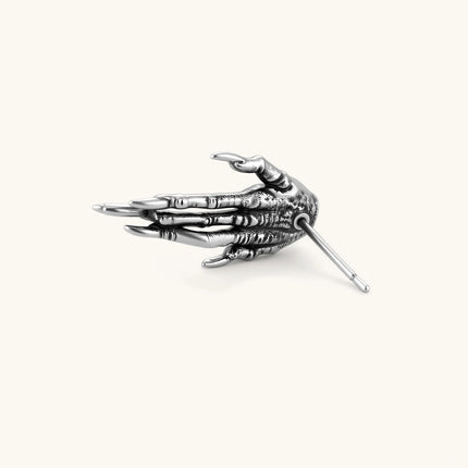 Silver stainless steel stud earring shaped like a skeletal demon claw hand with an eye motif on the palm and sharp talon fingers.